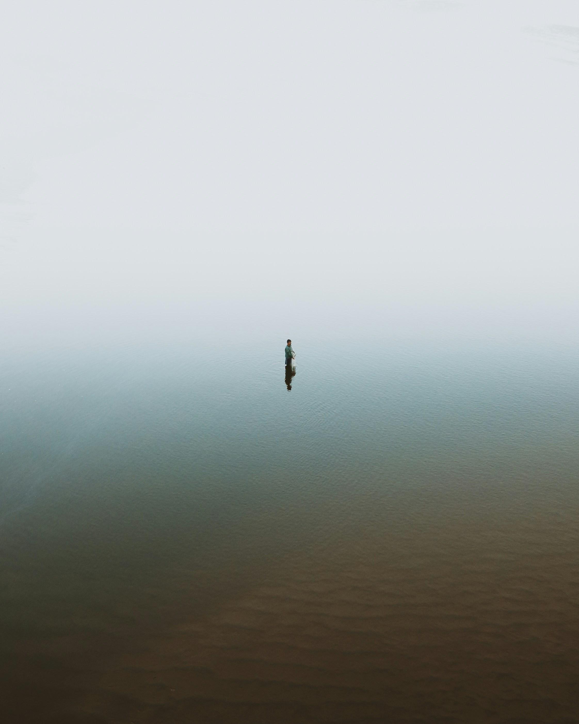 A lone fisherman stands waist-deep in a misty body of water during daytime, surrounded by fog, creating a serene and isolated atmosphere