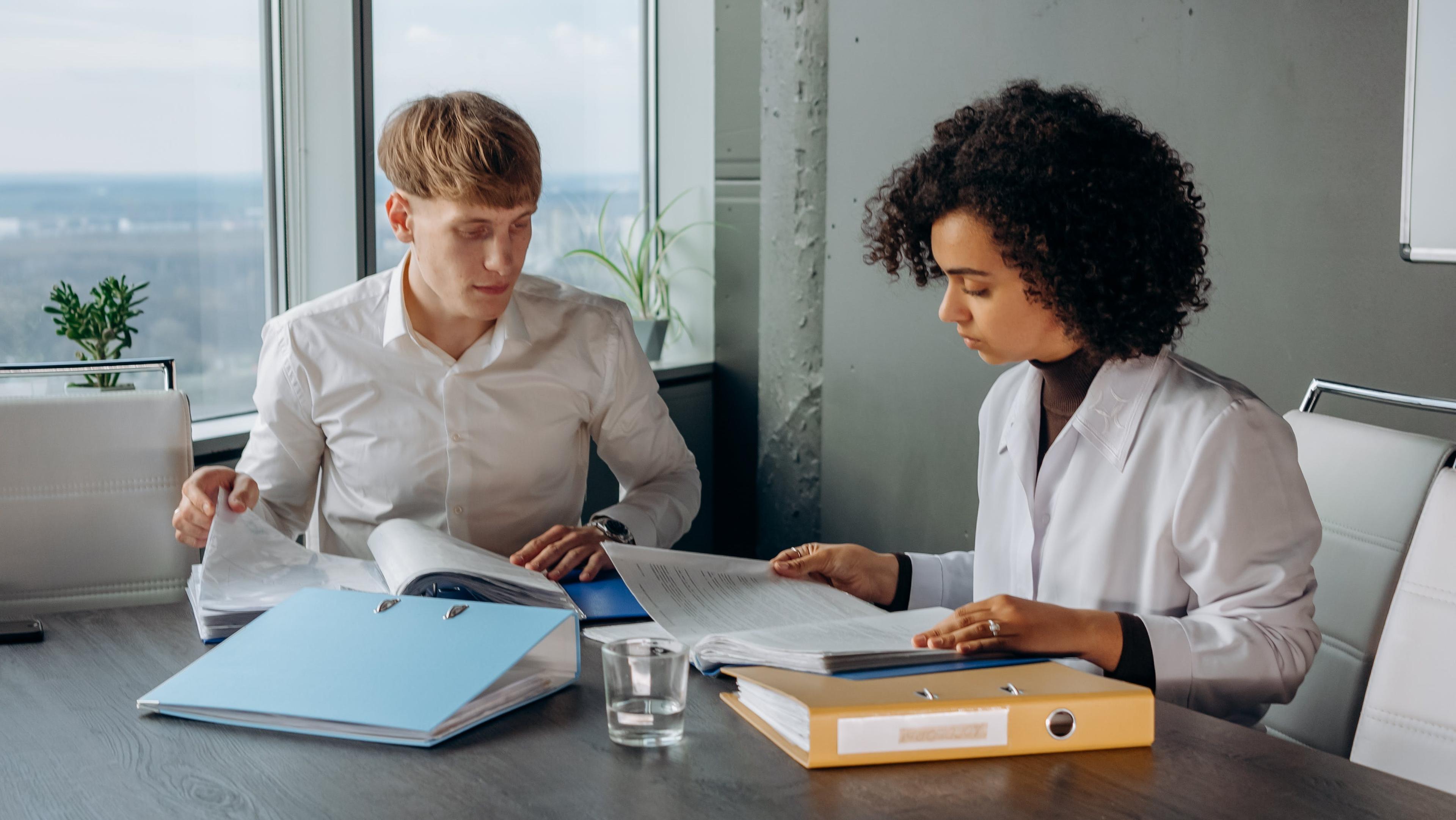 two people reading through books at a desk in an office