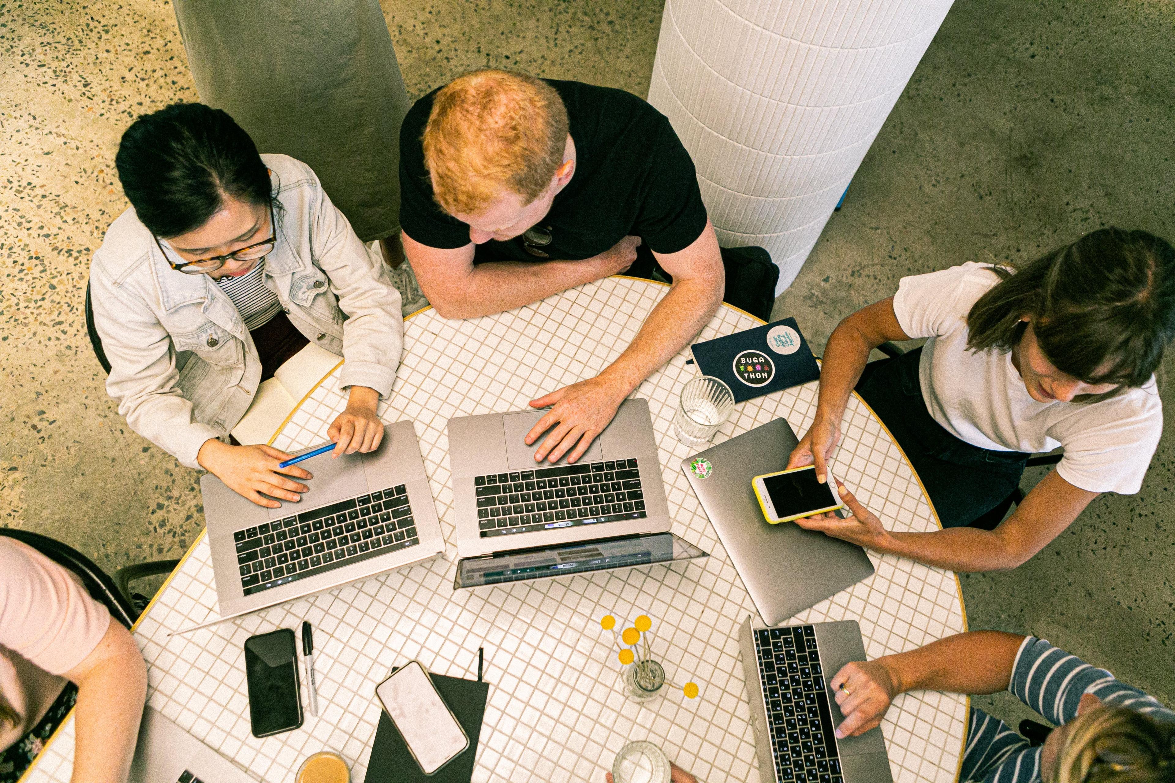 group of people sitting at a table with laptops, books and papers